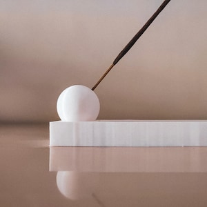 Könnte beinhalten: Weißer Keramik-Räucherstäbchenhalter mit runder Oberseite und einem langen, dünnen Räucherstäbchen, das darauf liegt.