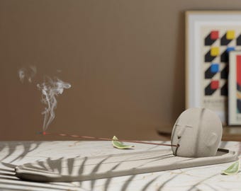 May include: A gray ceramic incense holder shaped like a face with a stick of incense burning and smoke rising from it. The incense holder is on a gray rectangular tray with a leaf on the tray.