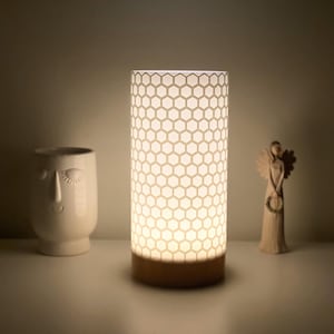 Puede incluir: Una lámpara de mesa blanca con una pantalla de patrón hexagonal y una base de madera. La lámpara está encendida e ilumina el área circundante.