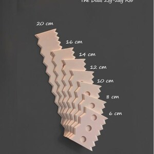 Puede incluir: Una pila de objetos en forma de zigzag de color rosa claro, cada uno con una longitud diferente, que oscila entre 6 cm y 20 cm. El texto "The Dual Zig-zag Rib" está en la parte superior.