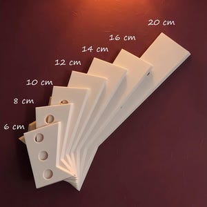 Peut inclure: Un ensemble d'étagères blanches et rectangulaires de différentes longueurs, de 6 cm à 20 cm, disposées en éventail. Les étagères sont fixées sur un mur rouge foncé. L'image comprend des mesures en centimètres.