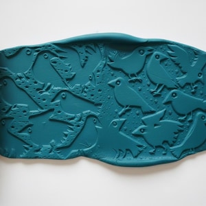 May include: A yellow rolling pin with a bird pattern next to a teal clay sheet with a bird design. The clay sheet has an embossed pattern of birds and stars. The rolling pin is cylindrical and has a repeating bird design.