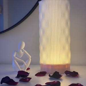 May include: A white ceramic lamp with a wavy, textured design. The lamp is lit and casting a warm glow. There are rose petals scattered on a white surface in front of the lamp.