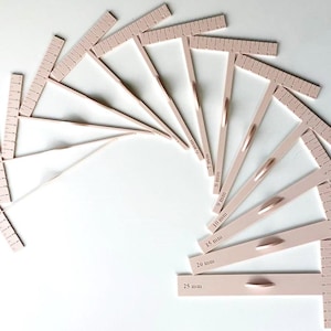 Op de afbeelding: Een waaier van zachtroze T-vierkanten, elk met een liniaal met millimeter markeringen en een handvat. De afmetingen zijn in millimeters aangegeven, waaronder 6 mm, 7 mm, 8 mm, 9 mm, 10 mm, 15 mm, 20 mm en 25 mm. De vierkanten liggen op een witte ondergrond.