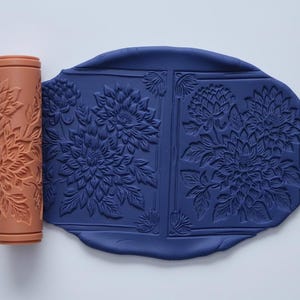 Könnte beinhalten: Ein terrakottafarbener Nudelholz mit floralem Muster neben einem Stück dunkelblauer Tonerde mit passendem floralem Design. Der Ton ist oval geformt und hat einen rechteckigen Rand mit dem floralen Design.