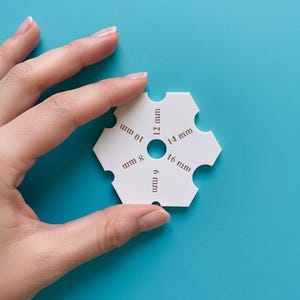 May include: A white, hexagonal gauge with cutouts and brown text indicating measurements from 6 mm to 16 mm. The tool is held against a bright blue background. The gauge is used for measuring.