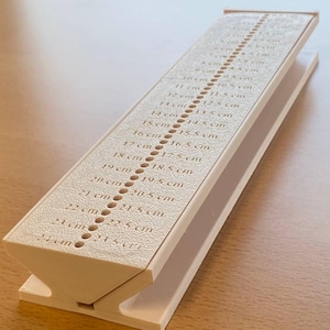 Op de afbeelding: Een beige plastic voetmeetapparaat. Het apparaat heeft een scharnierend ontwerp en is voorzien van een liniaal met metingen in centimeters, variërend van 8 cm tot 24 cm. De liniaal is gemarkeerd met stippellijnen en cijfers.