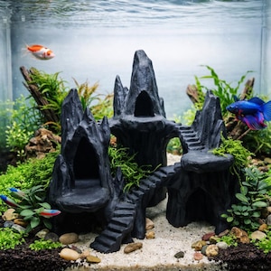 May include: A black aquarium decoration resembling a castle with multiple towers, archways, and staircases. The castle is surrounded by green plants, gravel, and sand. Several colorful fish swim in the water, creating a vibrant underwater scene.