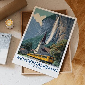 Peut inclure: Une affiche de voyage représentant un train jaune, une église et une cascade sur fond de montagnes. L'affiche porte l'inscription "WENGERNALPBANH SUISSE" en bleu. L'affiche est présentée sur un plateau en bois.