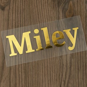 May include: Gold metallic name decal that reads "Miley" in a bold, sans-serif font. The letters are affixed to a clear, rectangular backing. The decal is displayed on a wooden surface.