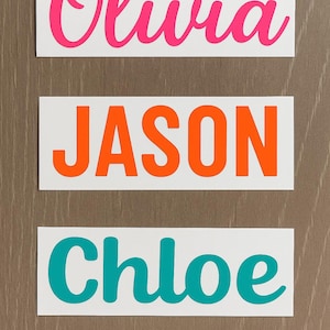 May include: Four white rectangular stickers with names in various colors. The names are Olivia in pink, Jason in orange, Chloe in teal, and Annabella in pink. The stickers are on a brown wooden surface.