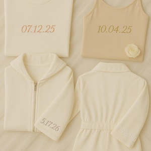 May include: Four ivory-colored garments are displayed: a crew neck t-shirt, a camisole, a zip-up hoodie, and a romper. Each item has a date embroidered in a light brown thread. A small white rose is attached to the camisole.