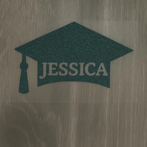 May include: A teal glitter graduation cap with the name "JESSICA" in a sans-serif font. The cap is on a clear rectangular background, set against a wood-grain surface. The tassel hangs on the left side of the cap.