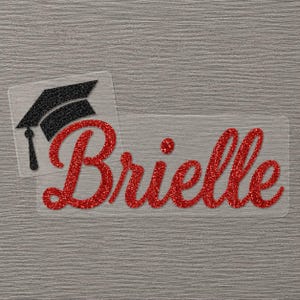 May include: A clear decal featuring the name Brielle written in a red glitter script font, accompanied by a black glitter graduation cap icon positioned to the left of the text. The design is presented on a transparent backing material.