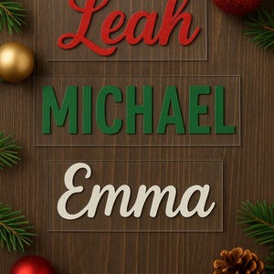 May include: Three name plaques with the names Leah, Michael, and Emma in red, green, and white lettering, respectively. The names are on clear acrylic rectangles, set against a dark wood background. Christmas ornaments and pine boughs complete the festive scene.