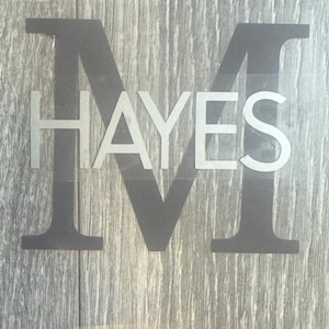 May include: A gray and white decal with the name "HAYES" layered over a large letter "M". The decal is on a wood-grain background. The letter "M" is a darker gray than the name "HAYES".