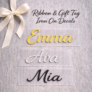 May include: Three iron-on decals with the names "Emma", "Ava", and "Mia" in gold, silver, and black glitter. The text "Ribbon & Gift Tag Iron On Decals" is above the names. A cream-colored ribbon is in the upper left corner.