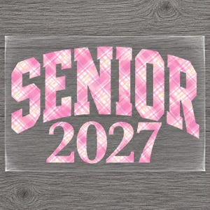 May include: A clear transfer sheet featuring the word SENIOR in a bold, arched block font, followed by the number 2027. The text has a pink, white, and yellow plaid pattern throughout, designed for application onto a garment or other surface.