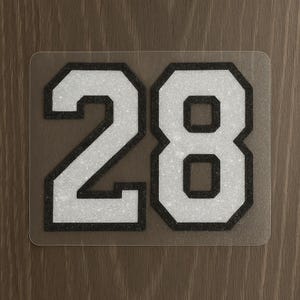 May include: A clear, rectangular sticker featuring the number "28". The number is white with a black outline and a textured, glittery appearance. The sticker is set against a brown background.