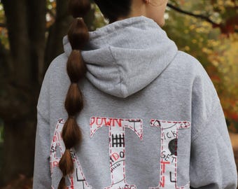 Puede incluir: Sudadera gris con capucha con un gran "ATI" bordado en rojo en la espalda. Las letras están rellenas de texto escrito a mano en blanco y negro.