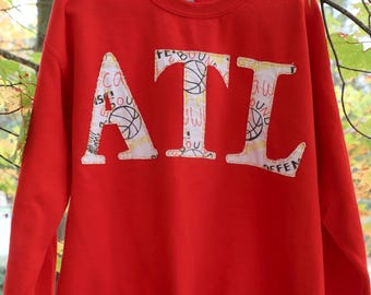 Puede incluir: Sudadera roja con un diseño blanco "ATL" hecho con recortes de tela con temática de baloncesto. Las letras están delineadas en negro.