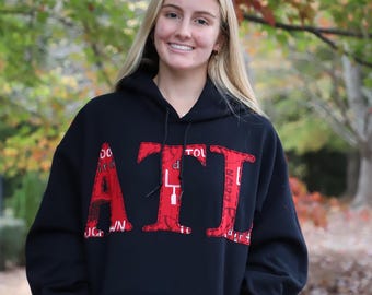 Puede incluir: Una sudadera con capucha negra con un gráfico rojo "ATL" en la parte delantera. Las letras están formadas por varios patrones rojos y blancos. La sudadera tiene una capucha con cordón y un bolsillo canguro.