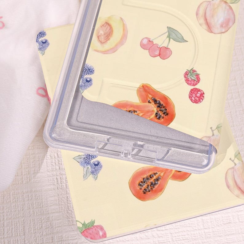 May include: A clear, rectangular phone case with a silver frame rests on a light yellow surface decorated with watercolor illustrations of fruits. The fruits include peaches, cherries, raspberries, papaya, and blueberries. The case is partially covering the fruit design.
