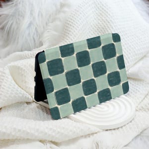 May include: A phone case with a green and white checkered pattern. The case is made of a hard plastic material and has a raised lip around the edges to protect the screen.