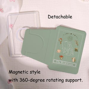 Könnte beinhalten: Ein magnetischer Handyhalter in Salbeigrün mit 360-Grad-Drehgelenk und abnehmbarem transparentem Rahmen. Die Oberfläche zeigt eine weiße Strichzeichnung einer lesenden Person, Sonne, Mondphasen, einen Pilz und den Schriftzug The Bookworm.
