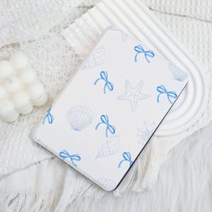 May include: A white case with a pattern of blue seashells, starfish, and bows. The case has a black border and is set against a white, textured background. The design has a nautical or beach theme.