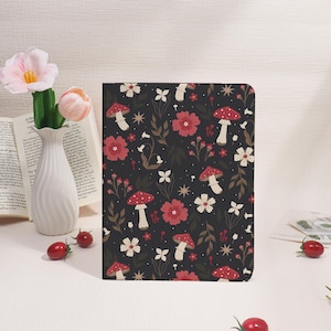 May include: A dark-colored journal with a repeating pattern of red mushrooms, flowers, and stars. The cover design features white and red flowers, brown leaves, and white mushrooms with red caps. The journal is rectangular and appears to be closed.