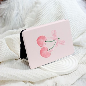 Może przedstawiać: Różowe etui na tablet z akwarelową ilustracją dwóch wiśni z różową wstążką. Etui leży na białej powierzchni.