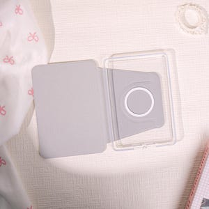 May include: A light gray tablet case, open to reveal a clear plastic frame and a gray interior with a white circular detail. The case is on a light-colored surface, with a white fabric with pink bows in the background.