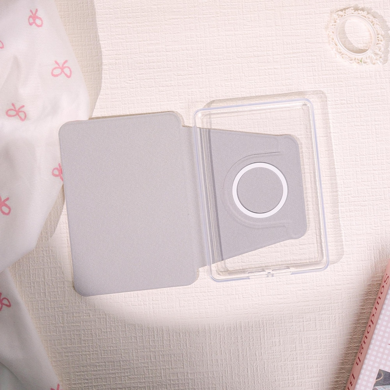 May include: A light gray tablet case, open to reveal a clear plastic frame and a gray interior with a white circular detail. The case is on a light-colored surface, with a white fabric with pink bows in the background.
