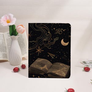 Pode incluir: Caderno preto com um design celestial dourado, com estrelas, uma lua crescente e notas musicais. As páginas abertas exibem notação musical. Um vaso branco com flores cor-de-rosa e um livro ao fundo.
