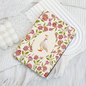 Puede incluir: Una funda rectangular con un diseño floral y un óvalo central con una ilustración de un ganso. La funda tiene un fondo crema con flores rosas y hojas verdes. El ganso tiene patas naranjas y un lazo morado.