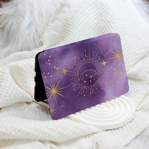 May include: Purple phone case with a gold crescent moon and stars design. The case has a watercolour effect and is on a white surface.