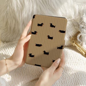 Peut inclure: Une housse de liseuse rectangulaire avec un fond marron clair à fines rayures verticales et un motif répétitif de petits chiens teckels noirs et feu. L'étui présente une finition lisse et recouvre la face avant de l'appareil électronique.