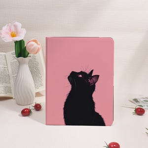 Può includere: Una custodia rosa per tablet con un'illustrazione di un gatto nero. Il gatto guarda in alto. La custodia ha una finitura liscia e uno sfondo rosa pieno.