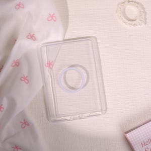 May include: A clear, rectangular phone case with a circular design in the center. The case is transparent, allowing the phone's color to show through. A white fabric with pink bow patterns is visible in the background.