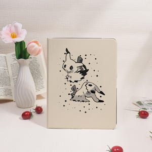 May include: A beige tablet case featuring a black and white illustration of two cartoon characters and scattered stars. The top character is upright, while the bottom one is lying down. The case is rectangular and has a smooth surface.