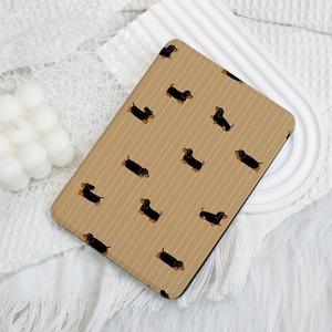 Pode incluir: Uma capa retangular com um padrão repetido de cães dachshund pretos com detalhes em castanho num fundo bege com riscas verticais. A capa está sobre uma superfície branca.