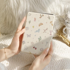 Può includere: Una custodia per e-reader color crema con un motivo ripetuto di piccole illustrazioni colorate, tra cui un croissant, un gatto, una farfalla, fragole, fiori, stelle, carte da gioco, una palla da discoteca e un bicchiere da cocktail.