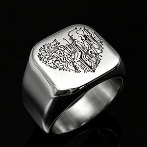 May include: A silver-toned metal signet ring featuring a flat, square face engraved with a black circuit board pattern arranged in the shape of a heart. The band has a smooth, polished finish and a wide, solid construction with a reflective surface.