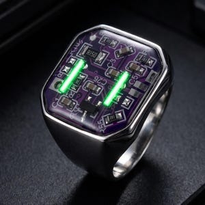 May include: A silver-toned ring featuring a rectangular face with a purple circuit board inlay. The inlay displays various electronic components, including resistors and capacitors, with two parallel glowing green light strips embedded in the center.