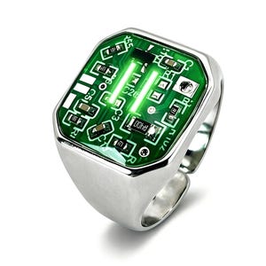 May include: A silver-toned ring featuring a rectangular face inlaid with a green circuit board pattern. The electronic design includes visible surface-mount components, white printed text, and metallic traces, set within a polished metal band.