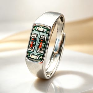 Puede incluir: Un anillo de tono plateado con una incrustación rectangular de una placa de circuito verde. La incrustación muestra dos pequeños componentes electrónicos naranjas colocados verticalmente en el centro de la placa verde, rodeados de circuitos plateados.