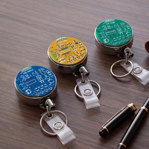 Può includere: Tre portabadge retrattili circolari con motivi a circuito stampato in blu, giallo e verde. Ogni portabadge presenta un involucro in metallo argentato, un anello metallico e un cinturino in plastica trasparente con bottone.