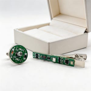 Puede incluir: Un gemelo circular y un pasador de corbata rectangular con placas de circuito impreso verdes y componentes electrónicos visibles, incluyendo resistencias y condensadores, montados sobre metal plateado, presentados con un joyero abierto.
