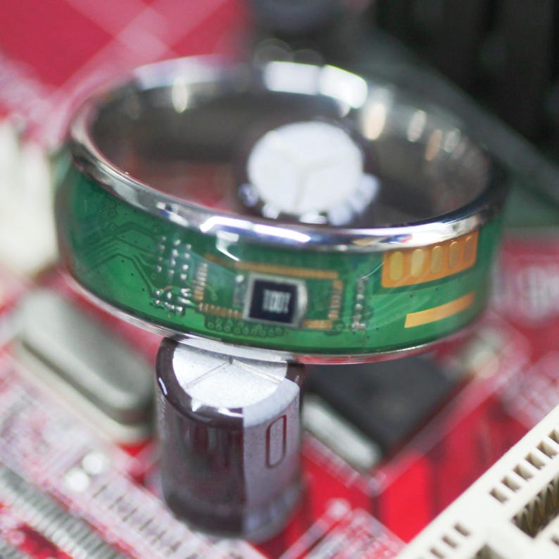 May include: A silver ring with a green circuit board design. The circuit board has a gold-colored chip and a black chip with the word "TODT" printed on it.